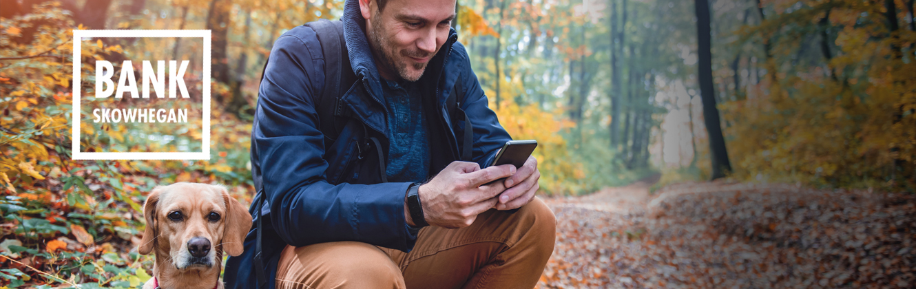 A man crouched over with his dog, while also looking at his phone